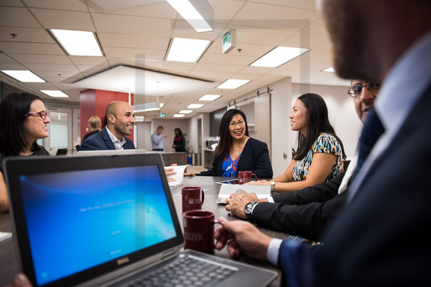 Photo de la vue par-dessus l’épaule d’un candidat au MBA pour cadres qui a son ordinateur ouvert et écoute cinq de ses collègues en pleine conversation; photo originale.